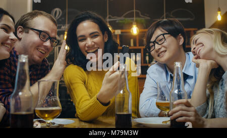 In the Bar/ Restaurant Hispanic Woman Makes Video Call with Her Friends. Group Beautiful Young People in Stylish Establishment. Stock Photo
