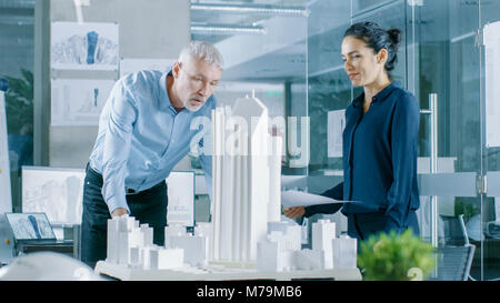 In the Architectural Bureau Female Engineer Brings Blueprints and Shows them to  Senior Architect, They Discuss Project. They Work as Urban Planners Stock Photo