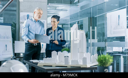 Male Senior Engineer Holds Laptop and has Discussion with Female Chief Engineer. They Work on a Building Model for a New District Urban Project Stock Photo
