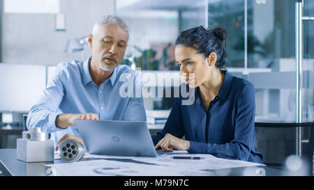 Experienced Male and Female Industrial Engineers Discuss ongoing Project while Working on a Laptop. They Design Machinery Component. Stock Photo