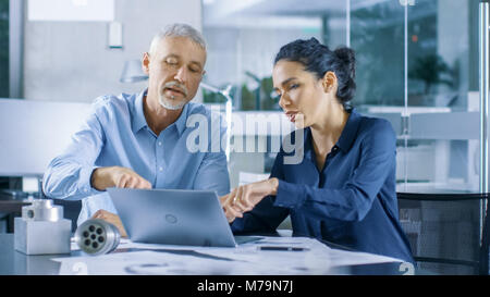 Experienced Male and Female Industrial Engineers Discuss ongoing Project while Working on a Laptop. They Design Machinery Component. Stock Photo