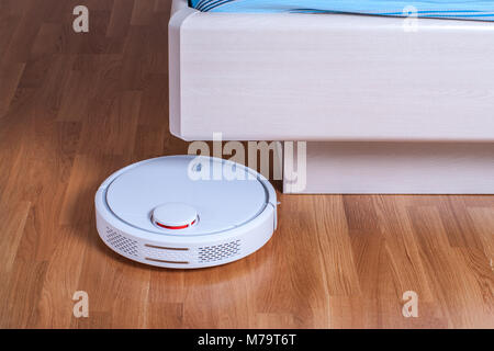 white Robot vacuum cleaner runs under bed in bedroom. Stock Photo