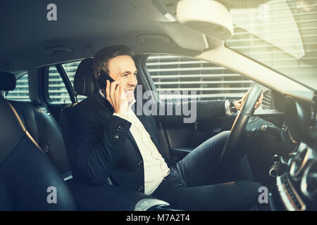 Smiling young businessman wearing a blazer talking on a cellphone while driving his car through the city during his morning commute to work Stock Photo