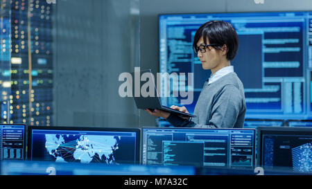 In the System Control Room IT Technician Works on a Laptop, in Background Multiple Displays with Graphics. Facility Works on Artificial Intelligence Stock Photo