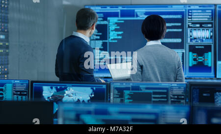 In the System Control Room IT Specialist and Project Engineer Have Discussion, they're surrounded by Multiple Monitors with Graphics. Stock Photo