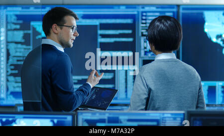In the System Control Room IT Specialist and Project Engineer Have Discussion while Holding Laptop, they're surrounded by Multiple Monitors Stock Photo