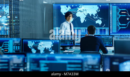 In the System Control Room Manager Holds a Briefing for His Staff Member. They're Work in Data Center and are Surrounded by Multiple Screens Stock Photo