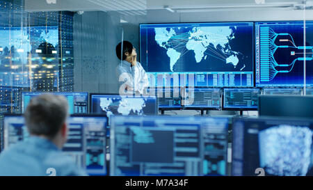 In the System Control Room Chief Engineer Thinks While Standing Before Big Screen with Interactive Map on it. Data Center is Full of Monitors Stock Photo