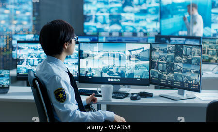 In the Security Control Room Officer Monitors Multiple Screens for Suspicious Activities, He Drinks from a Mug. He's Surrounded by Monitors Stock Photo