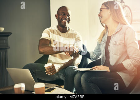 Two diverse young work colleagues sitting in a stylish modern office smiling and shaking hands together Stock Photo