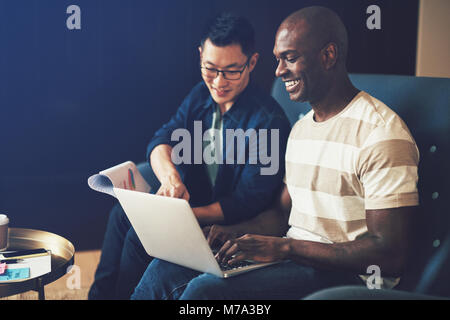 Two diverse work colleagues sitting together on a sofa in an office laughing while working on a laptop and discussing paperwork Stock Photo