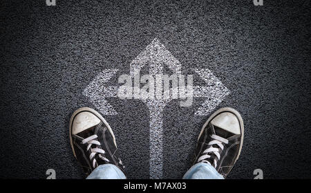 A teenager in jeans and canvas shoes standing on asphalt road with three-way arrow in the center and copy space. Concept of standing at the crossroads