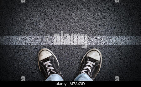 A teenager in jeans and canvas shoes standing on asphalt road behind a line with copy space. Concept of standing at a starting line, a new beginning, 