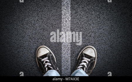 A teenager in jeans and canvas shoes standing on asphalt road with road divider marking in the center and copy space. Concept of standing at the cross