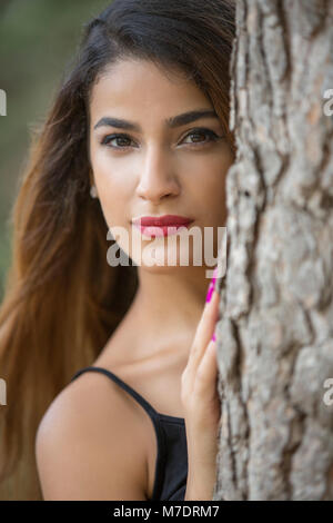 Beautiful young woman peeking from a behind a tree Stock Photo