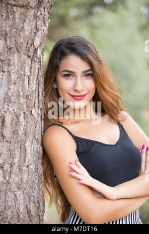 Happy young woman leaning against a tree arms folded smiling Stock Photo
