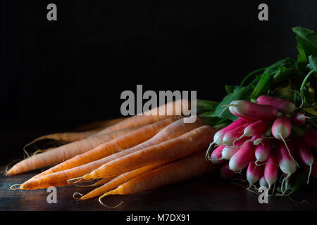A bunch of radishes and a fresh carrot bunch on a table with a black background in natural light. These vegetables are ingredients for many cooking re