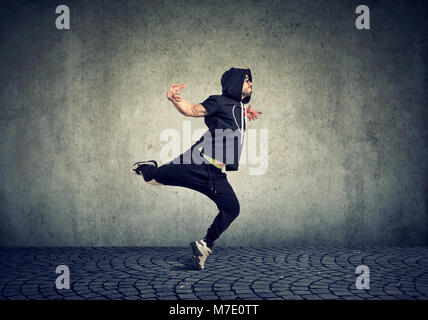 Stylish young man in trendy sportive clothing performing street dance with on gray wall background. Stock Photo