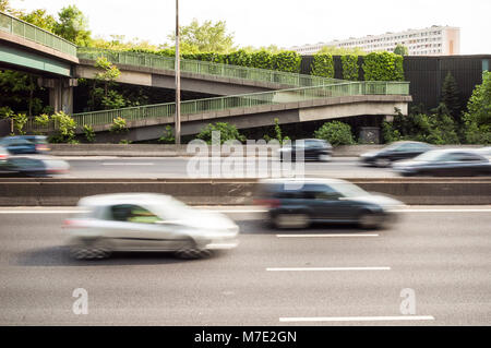 A highway with cars passing under a pedestrian footbridge, behind a noise barrier covered with vegetation, and residential building in the background. Stock Photo