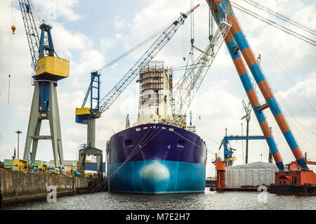 An old tanker moored in a dockyard, undergoing maintenance or repair, surrounded by large lifting cranes. Stock Photo