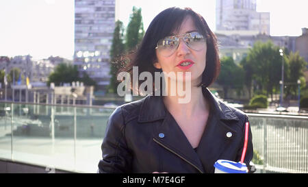 Beautiful brunette woman drinks from paper cup on a city urban surroundings with the sun backlight. Outdoor urban shot at sunset. The woman is wearing Stock Photo