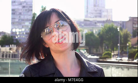 Beautiful brunette woman on a city urban surroundings with the sun backlight. Outdoor urban shot at sunset. The woman is wearing sunglasses and leathe Stock Photo