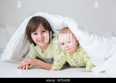 Cute little six month old baby boy and his older brother, playing under duvet at home in bed in bedroom, smiling happily Stock Photo