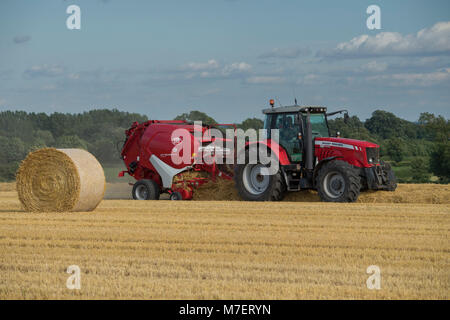 Baling straw in farm field, a farmer works & drives red tractor pulling round baler & passing large bale - Whixley, North Yorkshire, England, UK. Stock Photo