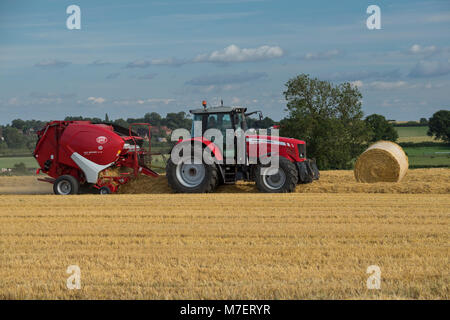 Baling straw in farm field, a farmer works & drives red tractor pulling round baler & passing large bale - Whixley, North Yorkshire, England, UK. Stock Photo