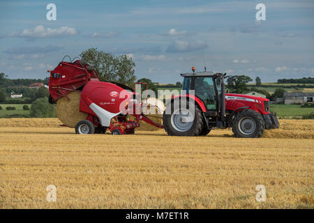 Baling straw in farm field, farmer works & drives red tractor pulling round baler (just releasing large bale) - Whixley, North Yorkshire, England, UK. Stock Photo