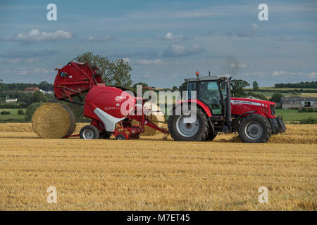 Baling straw in farm field, farmer works & drives red tractor pulling round baler (large bale just released) - Whixley, North Yorkshire, England, UK. Stock Photo