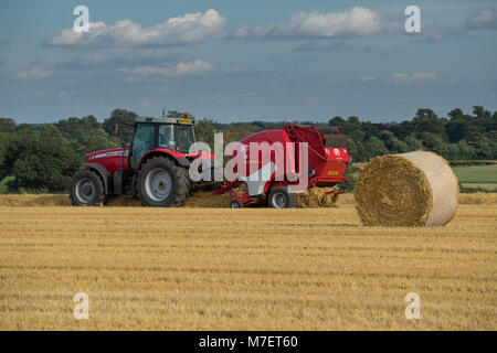 Baling straw in farm field, a farmer works & drives red tractor pulling round baler & passing large bale - Whixley, North Yorkshire, England, UK. Stock Photo