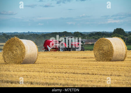 Farmer in red farm tractor pulling round baler, is driving & working in field, baling straw into round bales - Whixley, North Yorkshire, England, UK. Stock Photo