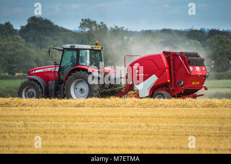 Farmer or man in bright red farm tractor pulling a round baler, is driving & working in a field, baling straw - Whixley, North Yorkshire, England, UK. Stock Photo