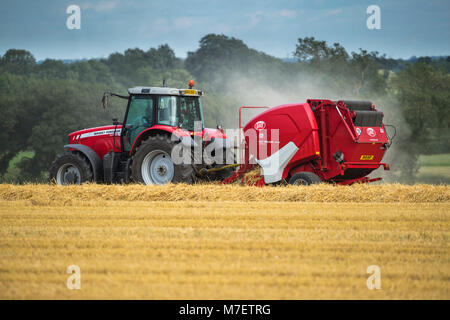 Farmer or man in bright red farm tractor pulling a round baler, is driving & working in a field, baling straw - Whixley, North Yorkshire, England, UK. Stock Photo