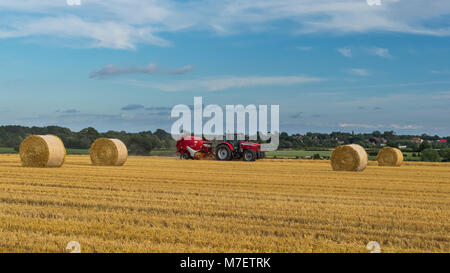 Farmer in red farm tractor pulling round baler, is driving & working in field, baling straw into round bales - Whixley, North Yorkshire, England, UK. Stock Photo