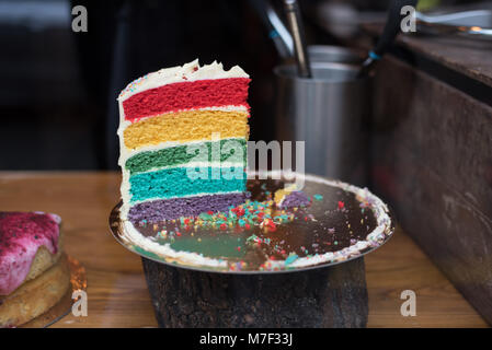 Yummy looking slice of rainbow sponge cake with white frosting on a cake stand with dark background Stock Photo