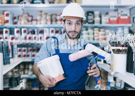 Positive workman in blue overalls with paint and brushes in hands in store Stock Photo