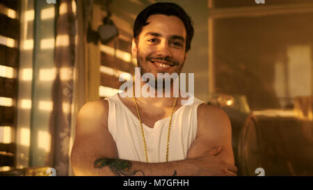 Stylish Hispanic Man Crosses Arms and Smirks/ Smiles into the Camera. In the Background Abandoned Building with Boarded up Windows. Stock Photo