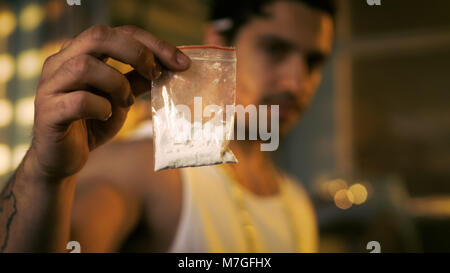 Brutal Drug Dealer Wearing Sleeveless Shirt and Gold Chain Holds and Offers Sample Bag Full of Drugs. He Lives in the Abandoned Building Stock Photo