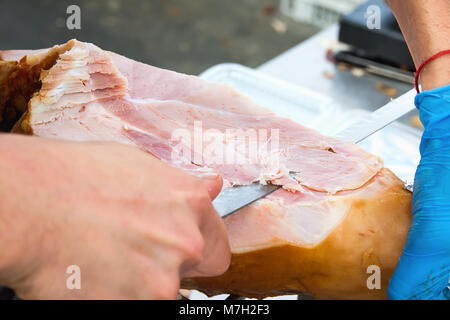 Farmer Slicing with Long Knife Smoked Baked Pork Ham from Leg at Market. Close up of Meat Texture. Vibrant Colors. Organic Produce. Travel Lifestyle.  Stock Photo