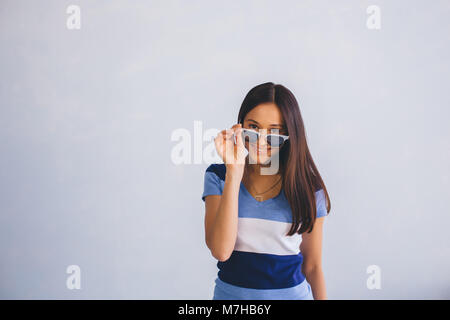 latino woman wearing casual clothes looking through sunglasses and smiling, while standing against background with copy space. Positive youth Stock Photo