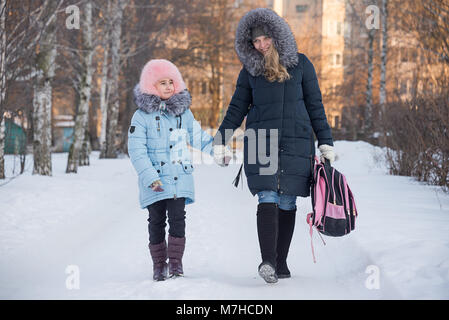 a young mother leads her daughter from school, the family smiles, children have fun, the family walks in the winter, go on the road. Stock Photo