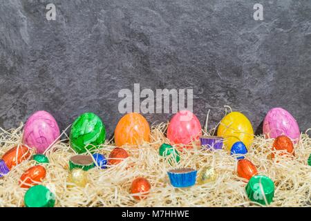 Colorful Easter eggs with chocolate easter eggs and sweet meat on hay in front of grey background Stock Photo