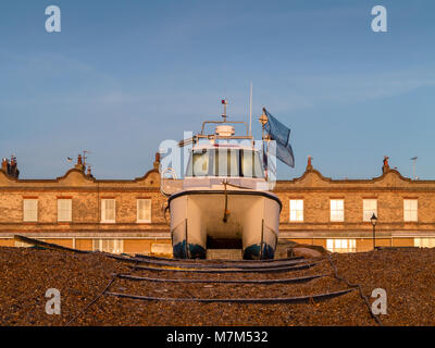 Front on symmetrical shot of a twin hull fishing boat moored on a bank of shingle in front of a building facade with evening light Stock Photo