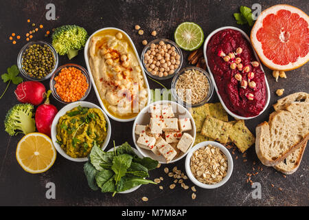 Vegan food background. Vegetarian snacks: hummus, beetroot hummus, green peas dip, vegetables, tofu. Top view, dark background, copy space. Stock Photo