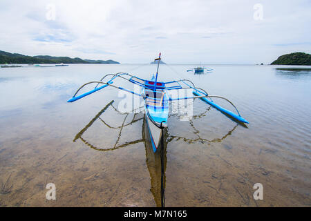 A traditional Filipino fishing boat with stabilising outriggers anchored in a perfectly still sea of a bay with small island on the horizon off of the Stock Photo