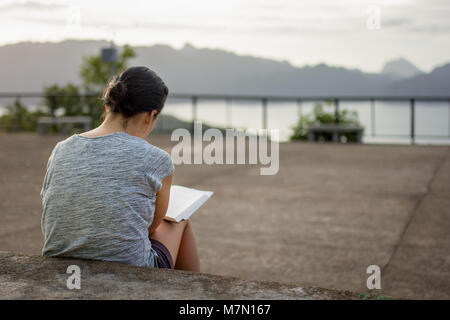 A young woman concentrates reading here hard back book whilst sat on some concrete steps. Stock Photo