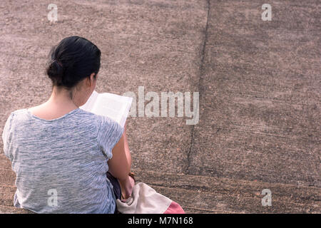 A young woman concentrates reading here hard back book whilst sat on some concrete steps. Stock Photo