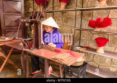 A Vietnamese woman wearing a traditional bamboo conical hat makes incense sticks using an old machine. Stock Photo